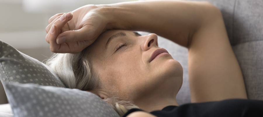 Yorgun ve halsiz görünen orta yaşlı bir kadın, koltuğa uzanmış şekilde gözleri kapalı, alnına eliyle dokunarak dinleniyor.