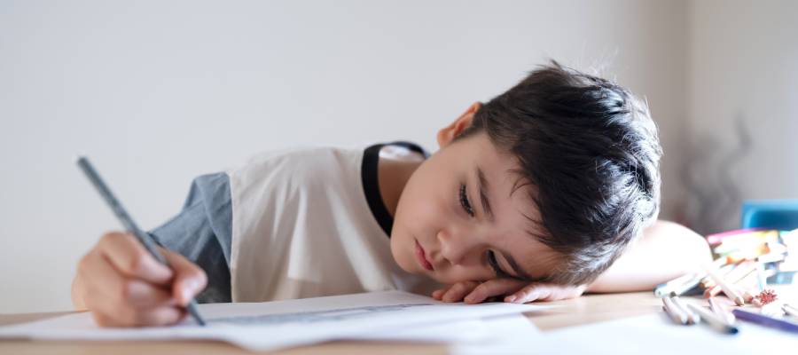 Başını masaya yaslamış, yorgun ve isteksiz görünen küçük bir çocuk; elinde kalemle ödev yapmaya çalışıyor. Yanında kırtasiye malzemeleri var.